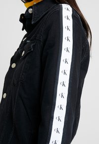 Chaqueta de mezclilla negra con herrajes plateados, una franja blanca prominente con el logo 'cK' repetido en la manga, y un diseño de cuello clásico.