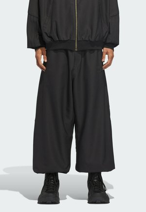 Hombre con pantalones negros de pierna ancha, chaqueta negra con cremallera y zapatillas negras, de pie contra un fondo claro y sencillo.