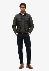 Schwarze Steppweste über einem grünen Strickpullover, dunkle Jeans und braune Bootsschuhe. Auffällige Details sind der geknöpfte Kragen und das dunkle Denim.
