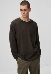 Koszula z długim rękawem w ciemnobrązowe paski z jasnobrązowymi pasami. Posiada kieszeń na piersi z subtelną naszywką z logo. Miękki materiał i luźny krój.