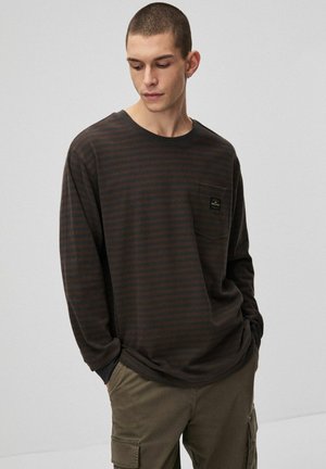 Koszula z długim rękawem w ciemnobrązowe paski z jasnobrązowymi pasami. Posiada kieszeń na piersi z subtelną naszywką z logo. Miękki materiał i luźny krój.