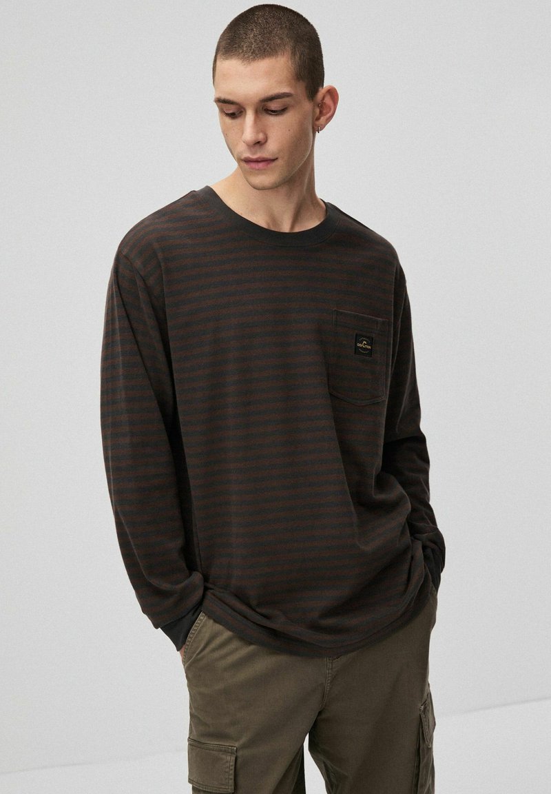 Koszula z długim rękawem w ciemnobrązowe paski z jasnobrązowymi pasami. Posiada kieszeń na piersi z subtelną naszywką z logo. Miękki materiał i luźny krój.
