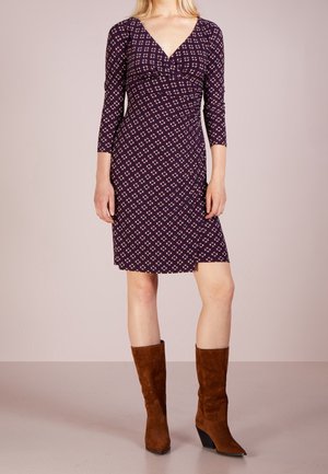 Femme portant une robe longue jusqu'aux genoux avec un motif sombre, des manches trois-quarts et des bottes en daim marron mi-mollet, debout devant un fond uni.