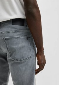 Grå denimjeans med en slät yta, med bakfickor och en svart läderlogga vid midjan. Handen vilar på låret.