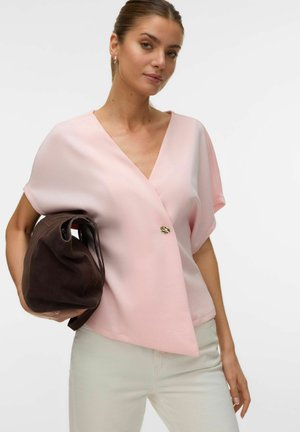 Kvinne iført en lyserosa kortermet bluse med en enkelt knapp, holder en stor mørk brun håndveske, stående mot en hvit bakgrunn.