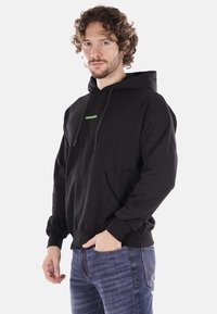 Felpa nera in cotone con tasca frontale, caratterizzata da un logo verde "PROPAGANDA" sul petto. Maniche lunghe e cappuccio regolabile.