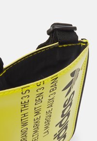 Gul syntetisk väska med svart text; har en svart tygfoder och sömmar. Justerbar rem och plastdetaljer synliga.