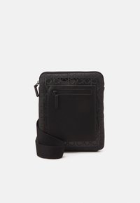 Czarna torba na ramię z wzorem logo Calvin Klein, z przednią kieszenią na zamek i regulowanym paskiem na ramię na białym tle.