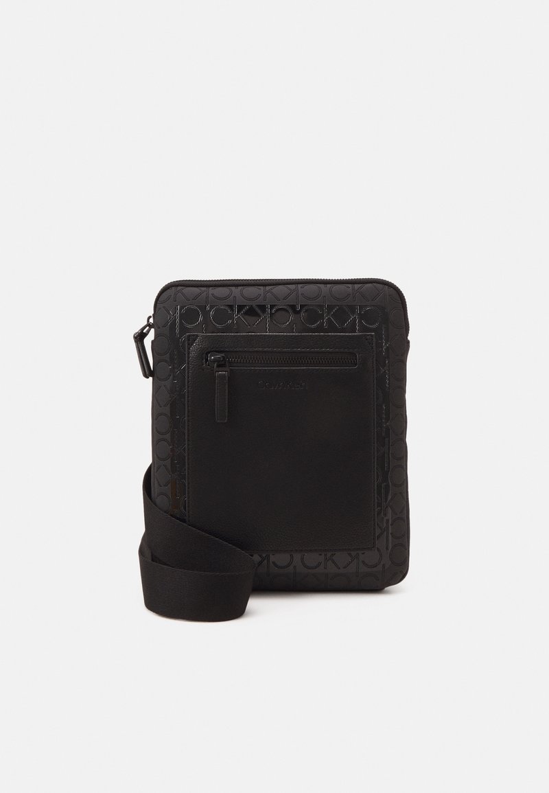 Czarna torba na ramię z wzorem logo Calvin Klein, z przednią kieszenią na zamek i regulowanym paskiem na ramię na białym tle.