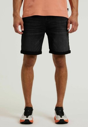 Mand iført sorte opsmåde denimshorts, ferskenfarvet t-shirt og hvide, sorte og orange sportssko, står opad mod en ensfarvet baggrund.