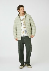 Lys grøn polstret jakke med hætte, båret over en tie-dye skjorte. Mørk grøn cargo-bukser med lommer, parret med grønne og cremede sneakers.