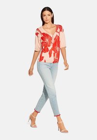 Helles rosa Sweatshirt mit rotem Blumenmuster, V-Ausschnitt und kurzen Ärmeln, kombiniert mit hellblauen Jeans mit roten Akzenten am Saum.
