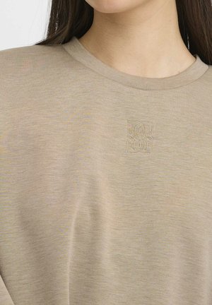 Beige kortærmet T-shirt lavet af blødt stof, med et diskret broderet logo i midten. Klassisk rund halsudskæring og afslappet pasform.