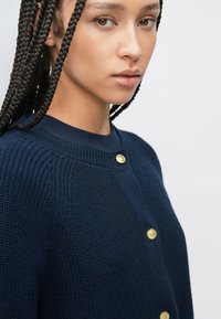 Μπλε σκούρο ζακέτα από υφή πλέξης, με στρογγυλή λαιμόκοψη και δύο χρυσά κουμπιά. Η κοντινή λήψη αναδεικνύει το μοτίβο της πλέξης.