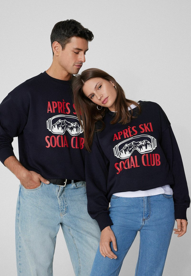 Navy Sweatshirts mit "APRÈS SKI SOCIAL CLUB" in rot bedruckt, mit einem grafischen Design von Skibrillen und Bergen. Über hellblauen Jeans getragen.