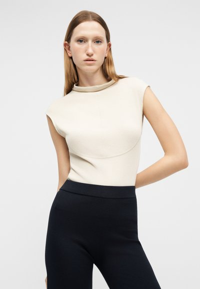 Γυναίκα με ίσια ανοιχτή καστανή τρίχα, ντυμένη με αμάνικο κρεμ τοπ και ψηλόμεσο μαύρο παντελόνι, στέκεται μπροστά σε απλό φόντο.