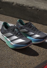 Pari mustavalkoisia Adidas juoksukenkiä, joissa vihreät ja siniset pohjat, asetettuna asfaltille lähelle katukiveä ja valkoista ajoradan viivaa.