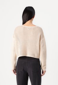 Cream-colored, cropped sweater with open-knit texture and long sleeves, paired with dark pants. Features a wide neckline and relaxed fit.