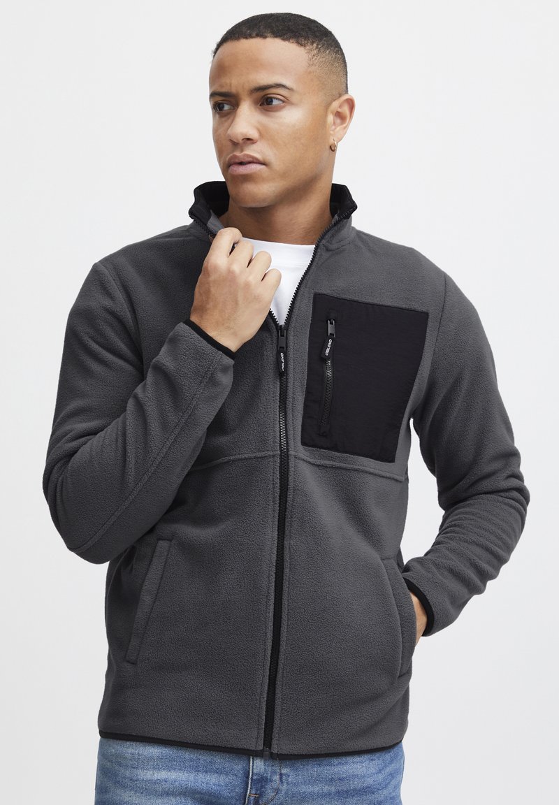 Chaqueta de forro polar gris con un bolsillo en el pecho negro, cierre delantero con cremallera, bolsillos laterales y un cuello alto. Presenta una textura suave y un corte casual.