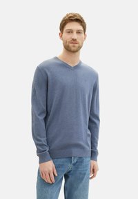 Maglione a V in morbido tessuto knit blu chiaro, maniche lunghe, polsini e orlo a costine, indossato con jeans blu chiaro. Dettaglio del logo semplice sul petto.