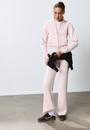 Femme portant un sweat à capuche rose clair et un pantalon, tenant un pull noir autour de sa taille, debout sur un sol gris avec un fond blanc.