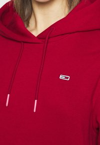 Sudadera roja con una textura suave, que cuenta con cordones elásticos con puntas rosas y un pequeño logo bordado en el pecho.