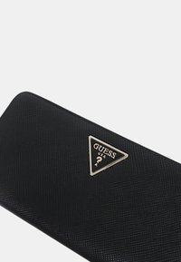 Portafoglio sintetico nero con superficie testurizzata. Presenta un logo triangolare dorato con "GUESS" e "USA" all'interno. Design compatto con angoli arrotondati.