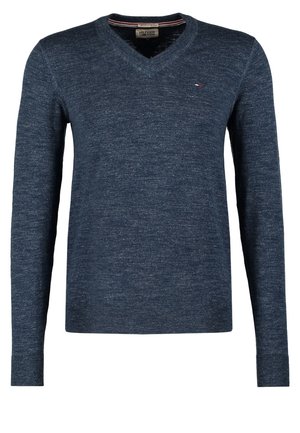 Uomo che indossa un maglione blu navy con scollo a V e pantaloni neri che tiene una borsa da viaggio nera con manici e dettagli in pelle marrone.