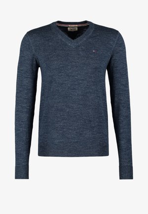 Mand iført marineblå V-hals sweater og sorte bukser, der holder en sort sportstaske med brune læderhåndtag og detaljer.