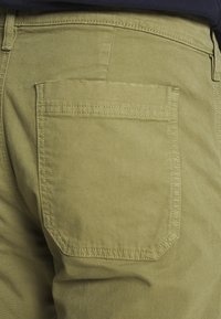 Close-up van de achterzak van khaki broek met zichtbare stiksels en een riemlus bij de taille.