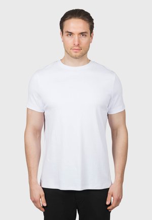 Mężczyzna o krótkich brązowych włosach, ubrany w gładki biały t-shirt z krótkim rękawem i czarne spodnie, stojący przodem na jasnoszarym tle.