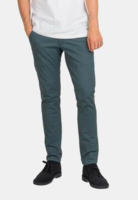 Pantalones chinos de algodón en color verde azulado oscuro, diseño ajustado, con bolsillos laterales y traseros, combinados con botas negras de cordones hasta el tobillo.