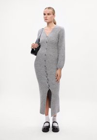 Abito grigio lavorato a maglia con scollatura a V, maniche lunghe e chiusura con bottoni frontale. Presenta una fessura alta fino alla coscia e una silhouette aderente.