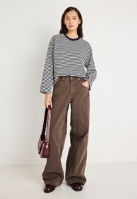 Ριγέ μπλούζα μακρυμάνικη σε ναυτικό μπλε και λευκό, συνδυασμένη με καφέ φαρδιά παντελόνια. Το μοντέλο κρατά μια μπορντό τσάντα με μακρύ λουράκι και φορά σκούρα παπούτσια.