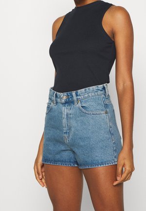Vrouw draagt een zwart geribbeld mouwloos topje en hooggetailleerde blauwe denimshorts, getoond van borst tot midden van het bovenbeen tegen een witte achtergrond.