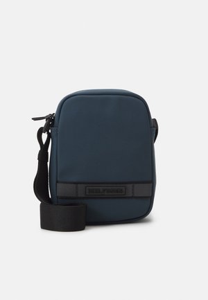 Petit sac bandoulière bleu marine avec bandoulière noire ajustable et détail patch noir horizontal à l'avant sur fond uni.