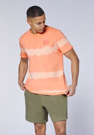 Mann trägt ein orange-weißes Batik-T-Shirt und olivgrüne Shorts, zieht am T-Shirt und schaut seitlich nach unten.