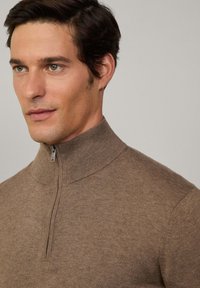 Smeđi zip-up džemper s visokim ovratnikom, napravljen od teksturirane tkanine. Ima metalni patentni zatvarač i uski kroj.