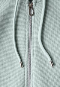 Felpa con zip di un verde chiaro tessurizzato, con zip argentata e dettagli metallici, dotata di cordino abbinato per la regolazione del cappuccio.