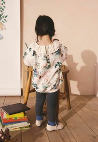 Pehmeä valkoinen paita, jossa on värikkäitä eläinkuvioita ja pitsikaulus, yhdistetty tummiin ribbineulottuihin leggingsseihin ja valkoisiin kenkiin. Lähistöllä on pino kirjoja.