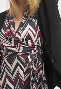 Šaty ve stylu wrap s chevron vzorem v burgundské, černé, krémové a šedé barvě. Měkká tkanina se zavazováním v pase, vrstvené pod černým kardigánem.