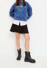 Sudadera azul con el texto "Bond Together" y un gráfico de cereza, superpuesta a una camiseta blanca. Falda negra plisada y botas de tobillo negras de plumas.