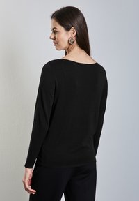 Donna con capelli lunghi e scuri, indossa una maglia nera a maniche lunghe e pantaloni, leggermente di lato, mostra la schiena e il profilo su sfondo semplice.