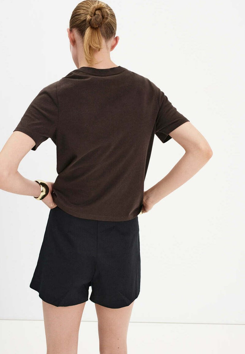 Femme aux cheveux blonds attachés en chignon, portant un t-shirt marron à manches courtes et un short noir, debout de dos, les mains sur les hanches.