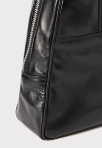 Close-up of shiny black leather bag corner with visible stitching and silver zipper detail on a light background.