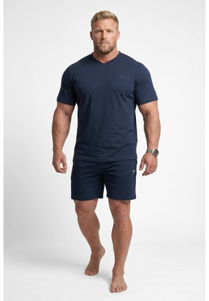 Muskulöser Mann, der barfuß geht, trägt ein eng anliegendes dunkelblaues T-Shirt und passende Shorts, mit einer Armbanduhr am linken Handgelenk.
