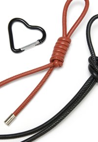 Cordino in pelle marrone con dettaglio a nodo, moschettone a forma di cuore nero e cordino intrecciato nero, che mettono in mostra texture e finiture variegate.