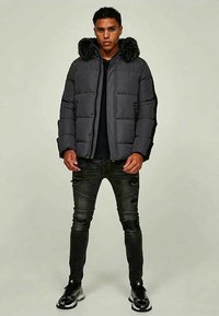 Man standing facing forward wearing a black puffer jacket with fur hood, ripped black skinny jeans, and black-and-white sneakers.