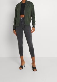 Groen gebreide ritscardigan, zwarte cropped top, hooggeknipte grijze spijkerbroek en zwarte open teen hakmuilen. Glad textuur, aansluitend ontwerp.