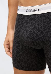 Detailní záběr na osobu, která má na sobě černé trenýrky Calvin Klein s opakujícím se vzorem loga a bílým elastickým pasem se značkou.